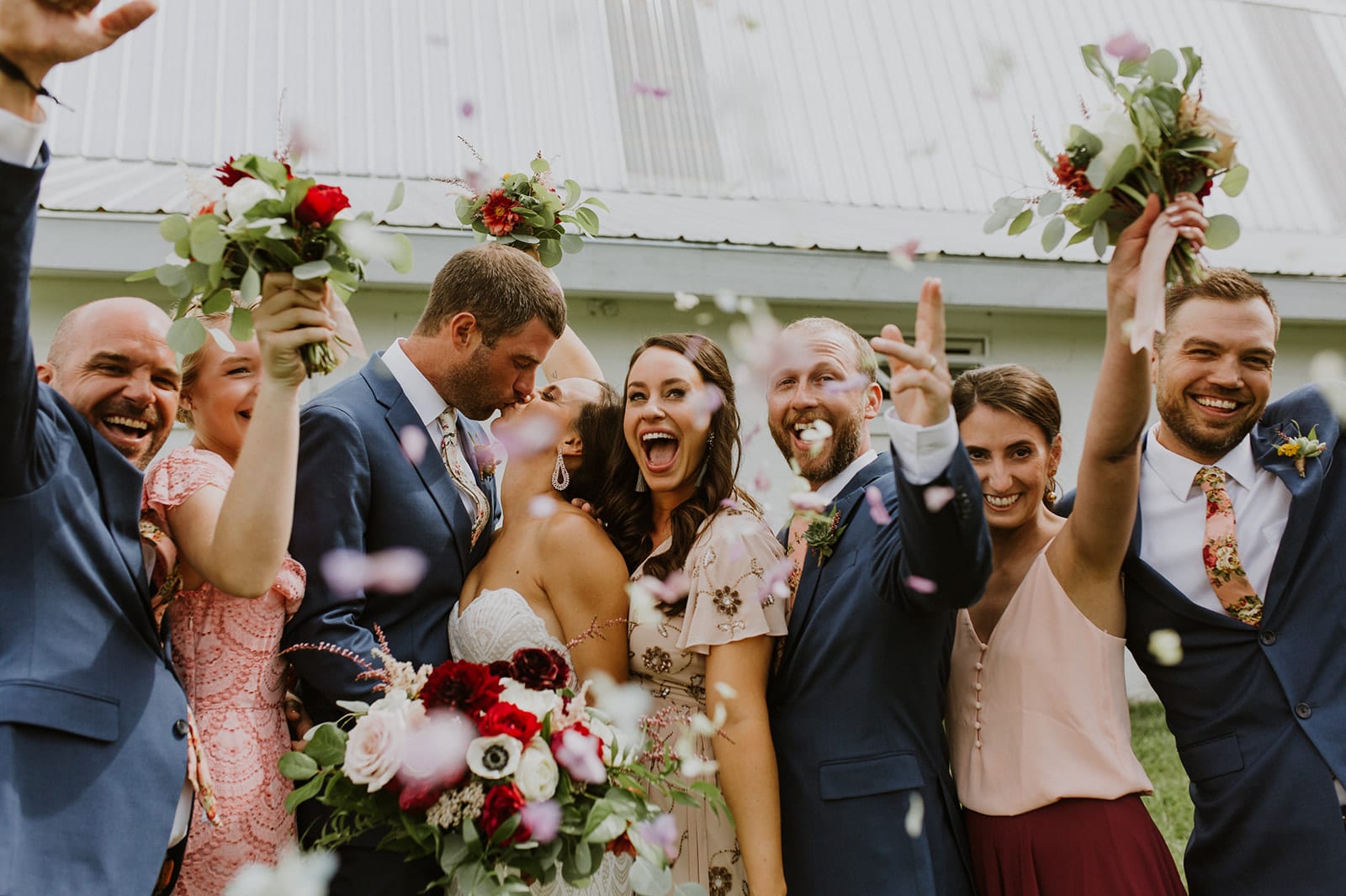 Celebratory bridal party, laughing and smiling while the happy bride and groom kiss