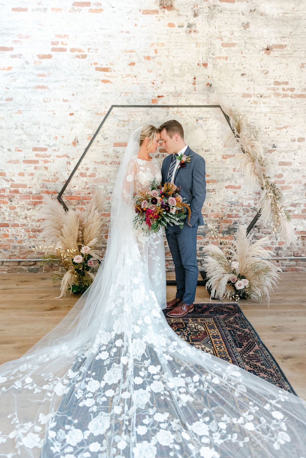 Bride and groom sharing a moment in front of their wedding arbor. The arbor is a metal octagon, and the background is brick. The bride is wearing a net dress with long veil and holding a colorful bouquet.