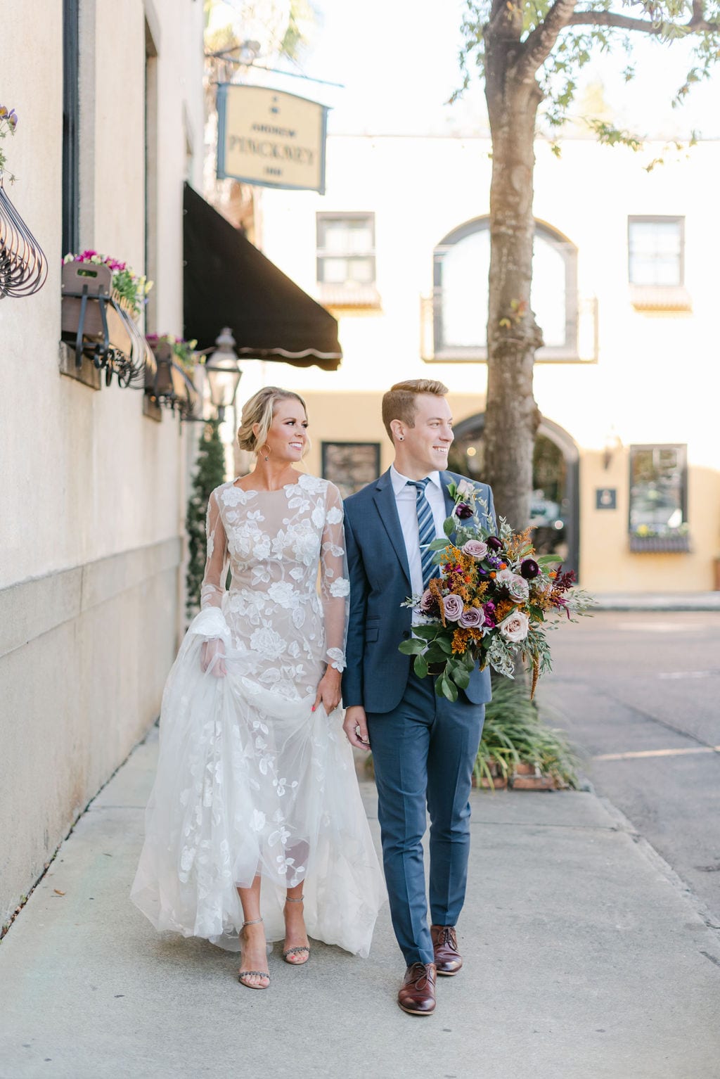 Bride and groom walking around downtown Charleston after their Charleston elopement. Bride is wearing a netted dress with floral accents, carrying and colorful bouquet.
